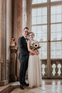 Ein Paar steht vor einem großen Fenster in einer eleganten Umgebung und fängt die Essenz einer Traumhochzeit ein. Der Mann trägt einen dunklen Anzug und die Frau ein weißes Kleid und hält einen Blumenstrauß in der Hand. Beide lächeln, und das natürliche Licht beleuchtet ihren besonderen Moment der Liebe und Freude.