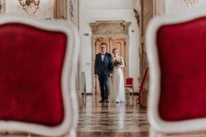 Ein Paar geht Hand in Hand einen großen Flur zwischen zwei roten Stühlen entlang, wobei die Frau einen Blumenstrauß hält. Die elegante Umgebung ist perfekt für eine Hochzeit, mit Marmorböden, Kronleuchtern und dekorativen Wänden, die zeitlose Romantik und sorgfältige Hochzeitsplanung widerspiegeln.