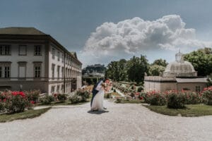 Ein Paar küsst sich romantisch in einem Garten, umgeben von blühenden roten Blumen, perfekt für diejenigen, die von ihrer Hochzeit träumen. Links steht ein großes Gebäude mit kunstvollen architektonischen Details auf der rechten Seite, während flauschige Wolken über den hellen Himmel ziehen.