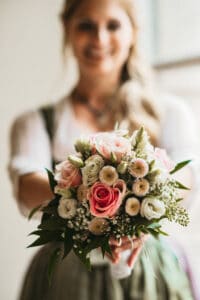 Eine lächelnde Frau in traditioneller Kleidung, vielleicht Teil ihrer Hochzeitsplanung, hält inmitten üppigen Grüns einen Strauß leuchtend rosa und weißer Rosen in die Höhe. Der Fokus liegt auf den atemberaubenden Blumen, die die Freude und Schönheit dieses Heiratsmoments vor einem sanft verschwommenen Hintergrund hervorheben.