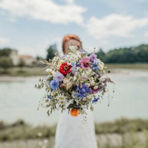 Eine Braut steht an einem ruhigen Fluss und hält einen großen Strauß Wildblumen in der Hand, der ihr Gesicht verdeckt. Die leuchtenden Blumen symbolisieren den freudigen Geist ihrer Hochzeit vor einem Hintergrund aus leicht verschwommenen Bäumen und einem bewölkten Himmel.