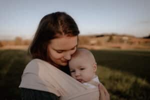 Eine Frau hält sanft ein Baby in einer weichen Trage und steht auf einem offenen Feld mit dunstigem, warm getöntem Hintergrund. Beide haben einen ruhigen Gesichtsausdruck, als würden sie die Ruhe dieses Augenblicks genießen, ganz so, als würden sie die Ruhe vor einer Hochzeit genießen.