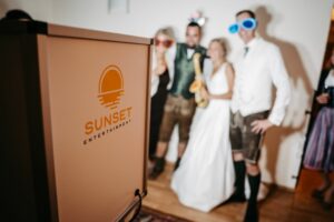 Eine Gruppe von vier Personen in festlicher Kleidung posiert für ein Foto im Innenbereich. Sie tragen helle, farbenfrohe Sonnenbrillen und lächeln neben einer Fotokabine mit dem Logo „SUNSET ENTERTAINMENT“ – perfekt, um Erinnerungen an Ihren besonderen Hochzeitstag festzuhalten!