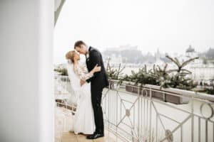 Braut und Bräutigam umarmen sich auf einem Balkon mit Blick auf eine Stadtlandschaft mit historischen Gebäuden und einem Schloss, perfekt für ihre Hochzeit. Die Braut trägt ein langärmeliges Spitzenkleid, der Bräutigam einen dunklen Anzug. Immergrüne Pflanzen schmücken an diesem bewölkten Tag das Geländer und symbolisieren zeitlose Liebe.