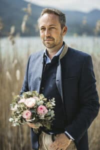 Ein Mann in einer traditionellen dunklen Jacke steht draußen und hält einen Strauß rosa Rosen und Grünzeug in der Hand, was die Essenz einer Hochzeit verkörpert. Hohes Schilf und eine verschwommene Landschaft mit Bergen und Wasser bilden die perfekte Kulisse für diesen Moment der Hochzeitsplanung.