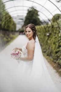 Eine Braut in einem weißen Hochzeitskleid und Schleier lächelt, während sie einen Strauß rosafarbener und weißer Blumen hält. Sie steht auf einem von Grün und einer tunnelartigen Struktur umgebenen Weg und fängt die bezaubernde Essenz einer Hochzeit ein.