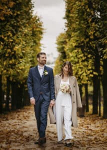 Ein Paar geht Hand in Hand einen von Bäumen gesäumten Weg entlang, auf dem Herbstlaub liegt. Die Umgebung ist perfekt zum Heiraten. Der Mann trägt einen dunklen Anzug und die Frau, in einem beigen Mantel mit einem Blumenstrauß in der Hand, träumt von Hochzeitsplänen. Die Szene ist heiter und romantisch.