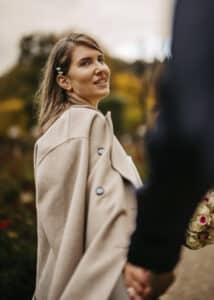 Eine Frau mit langen Haaren und einem hellbeigen Mantel blickt zurück, während sie die Hand einer anderen Person, vielleicht ihres Partners, hält. Sie steht im Freien vor verschwommenem Grün und suggeriert damit eine Hochzeit in der Natur. Ein Blumenstrauß lässt auf romantische Heiratsmomente schließen.