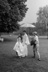 Ein Paar in Hochzeitskleidung lächelt unter einem Regenschirm, den eine Person für sie hält. Sie gehen über eine Wiese mit Bäumen im Hintergrund. Die in Schwarzweiß festgehaltene Szene spiegelt wunderschön einen ungezwungenen Moment der Hochzeitsfreude wider.