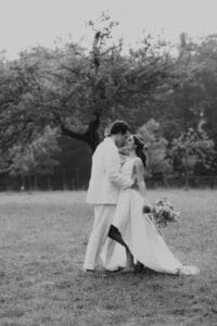 Ein Paar in weißer Hochzeitskleidung umarmt sich auf einer Wiese und verkörpert den Geist der Hochzeit. Die Braut steht mit ihrem Brautstrauß in der Hand neben ihrem Bräutigam unter einem großen Baum. Dieser zeitlose Moment der Liebe und Hingabe ist wunderschön in Schwarzweiß festgehalten.