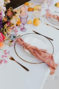 Ein wunderschön gedeckter Tisch, perfekt für eine Hochzeit, mit einem Blumenschmuck, bunten Kerzen und Zitronen. Eine rosa Stoffserviette ist kunstvoll auf einem weißen Teller mit Goldrand befestigt. Rosa Gläser und goldenes Besteck werten das elegante Ambiente noch weiter auf, ideal für Ihren besonderen Tag.