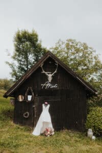 Eine rustikale Scheune aus Holz steht auf einer grünen Wiese. An der Tür hängt ein Brautkleid, das den Charme der Hochzeitseleganz verkörpert. An der Scheune sind Geweihe und dekorative Spiegel angebracht. Am Sockel ruht ein Blumenstrauß, über dem der Name „Miri“ steht.