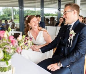 Braut und Bräutigam sitzen während ihrer Hochzeitszeremonie an einem Tisch, halten Händchen und lächeln freudig. Die Braut trägt ein weißes Kleid mit einem Blumenstrauß, während der Bräutigam einen Anzug mit Blumen am Revers trägt. Die Gäste sitzen im Hintergrund und werden Zeuge dieses wunderschönen Heiratsmoments.