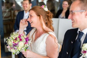 Eine lächelnde Braut mit roten Haaren, gefangen in den Emotionen ihres Hochzeitstages, hält einen Strauß weißer und violetter Blumen in der Hand. Sie wischt sich eine Träne aus dem Auge, während sie neben einem Mann im Anzug sitzt, umgeben von anderen glücklichen Gästen.