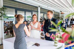 Ein Paar steht lächelnd bei seiner Hochzeitszeremonie und steht einer Frau gegenüber, die die Trauung durchführt. Die Braut trägt ein weißes Kleid und der Bräutigam einen Anzug. Im Hintergrund sitzen die Gäste und genießen das wunderschön geplante Ereignis. Eine Vase mit Blumen schmückt den Tisch davor.