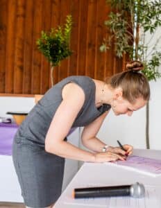 Eine Frau in einem grauen Kleid unterschreibt ein Dokument bei etwas, das ihre Hochzeit zu sein scheint. Im Vordergrund steht ein Mikrofon. Holzvertäfelungen und eine Topfpflanze rahmen diesen intimen Moment der Hochzeitsplanung elegant ein.