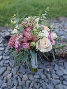 Ein Strauß verschiedener Blumen, darunter rosa Proteas, cremefarbene Rosen und Eukalyptusblätter, wird auf einem Bett aus grauen Steinen mit grünem Gras im Hintergrund platziert – perfekt für die Kulisse einer Hochzeit oder als Inspiration für diejenigen, die sich intensiv mit der Hochzeitsplanung beschäftigen.