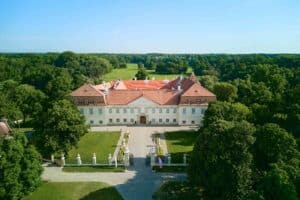 Luftaufnahme eines großen, weißen historischen Herrenhauses, das sich perfekt für die Hochzeitsplanung eignet und von üppigen grünen Bäumen umgeben ist. Das Gebäude verfügt über ein rotes Ziegeldach und ein symmetrisches Design. Ein Kiesweg führt zu seinem bezaubernden Eingang. Der Himmel ist klar und blau, eine ideale Kulisse für eine Traumhochzeit.