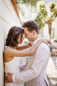 Ein Paar umarmt sich, die Stirnen berühren sich, die Augen sind geschlossen und es lächelt. Die Frau hält einen Blumenstrauß. Beide sind für ihre Hochzeit in elegante Hochzeitskleidung gekleidet und stehen vor einer sonnenbeschienenen Wand mit Bäumen im Hintergrund.