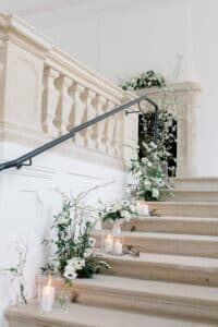 Eine mit Kerzen und weißen Blumen dekorierte Steintreppe schafft die Kulisse für eine Traumhochzeit. Das schlichte Geländer und die Blumenarrangements mit Grünpflanzen säumen elegant die Stufen und bieten eine helle, minimalistische Kulisse, die perfekt für jede Hochzeitsplanung ist.