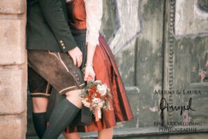 Ein Paar in traditioneller Kleidung steht dicht beieinander und verkörpert die Essenz einer Hochzeit. Die Frau hält einen Blumenstrauß. Auf einer strukturierten Wand im Hintergrund sind die Texte „Mario & Laura“ und „Fine Art Wedding Photographie“ zu sehen, die einen Moment der Eleganz und Liebe einfangen.