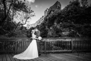 Ein Schwarzweißfoto zeigt ein Paar, das sich in Hochzeitskleidung auf einer Holzbrücke umarmt, eingerahmt von einem üppigen, baumbedeckten Hügel und einem historischen Gebäude. Die Braut steht mit ihrem Brautstrauß unter einem teilweise bewölkten Himmel – ein perfekter Moment des Hochzeitsglücks.