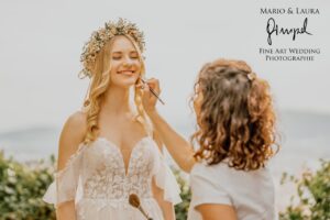 Eine Braut mit Blumenkrone lächelt, während eine Visagistin ihr Make-up aufträgt. Sie sind an einem sonnigen Tag im Freien, umgeben von Grün, und feiern ihre Hochzeit. Das Bild enthält den Text: „Mario & Laura Gumpel Fine Art Hochzeitsfotografie.