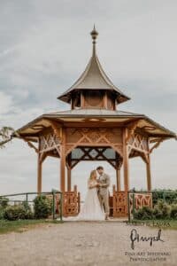 Ein Paar steht vor einem verzierten hölzernen Pavillon unter einem bewölkten Himmel. Die Frau trägt ein wallendes Kleid und der Mann einen Anzug und hält Händchen. Die Szene ist von Grün umgeben – ein perfekter Moment für diejenigen, die ihre Hochzeit planen. In der Ecke verewigt „Fine Art Wedding Photographie“ diese elegante Erinnerung.