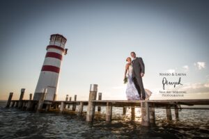 Braut und Bräutigam stehen bei Sonnenuntergang auf einem Holzsteg neben einem Leuchtturm und fangen die Essenz ihrer Hochzeit ein. Sie trägt ein weißes Kleid und hält Blumen in der Hand, er einen Anzug. Der Himmel ist klar und das Wasser plätschert sanft unter ihnen – ein perfekter Moment in ihrer Hochzeitsplanung.