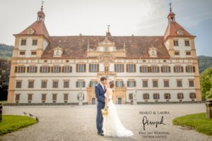 Braut und Bräutigam küssen sich vor einem prachtvollen, historischen Gebäude mit kunstvoller Architektur, das die Essenz ihrer Hochzeit verkörpert. Die Braut hält einen Blumenstrauß und der Bräutigam trägt einen Anzug. Es ist ein sonniger Tag, der das rote Dach und die zahlreichen Fenster dieses traumhaften Hochzeitsortes in Szene setzt.