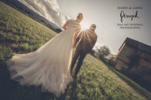 Braut und Bräutigam gehen bei Sonnenuntergang Hand in Hand über eine Wiese und verkörpern damit die Essenz einer Hochzeit. Die Braut trägt ein fließendes weißes Kleid, während die Sonne durch die Wolken lugt und eine warme, leuchtende Kulisse schafft. In der Ferne steht eine alte Scheune, die ihrer Hochzeitskulisse rustikalen Charme verleiht.