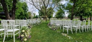 Die bezaubernde Anordnung der Hochzeitszeremonie im Freien besteht aus Reihen weißer Stühle vor einer mit Blumen geschmückten Laube. Umgeben von üppigen grünen Bäumen führt ein mit Blumenarrangements gesäumter Weg zur Laube und macht sie zu einem perfekten Zufluchtsort für diejenigen, die mit Stil heiraten.