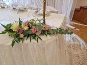 Ein Blumenarrangement mit rosa, weißen und violetten Rosen, Grünpflanzen und Eukalyptusblättern schmückt einen mit weißer Spitze bedeckten Tisch – eine perfekte Note für eine Hochzeit. Gläser und Geschirr sind elegant im Hintergrund arrangiert.