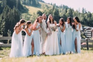 Eine Braut und ein Bräutigam stehen lächelnd mit einer Gruppe von sieben Brautjungfern in hellblauen Kleidern während ihrer Hochzeit. Sie sind im Freien, umgeben von einer malerischen Kulisse aus Bäumen und Hügeln. Alle wirken fröhlich, die Brautjungfern klatschen und jubeln.