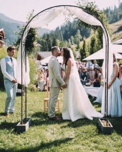 Braut und Bräutigam stehen sich während ihrer Hochzeit unter einem dekorierten Bogen gegenüber. Die Gäste sitzen auf beiden Seiten, mit einer malerischen Kulisse aus Bäumen und Bergen, die das Ergebnis einer sorgfältigen Hochzeitsplanung widerspiegelt. Eine Person in einem hellen Anzug steht seitlich.