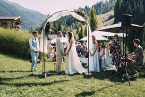 Vor einer atemberaubenden Bergkulisse findet die Hochzeit des Paares unter einem bezaubernden Bogen statt. Brautjungfern in hellblauen Kleidern und Gäste sitzen, während Musiker mit Gitarre und Keyboard in der Nähe spielen. Die idyllische Umgebung ist üppig und sonnendurchflutet, perfekt für diese Hochzeitsfeier im Freien.