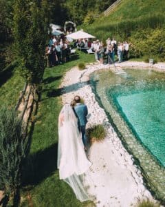 Braut und Bräutigam gehen einen Weg neben einem klaren Teich entlang und nähern sich den draußen versammelten Gästen. Mit ihrem langen wehenden Schleier verkörpert die Braut an diesem sonnigen Tag ihrer Hochzeit Eleganz inmitten des Grüns.