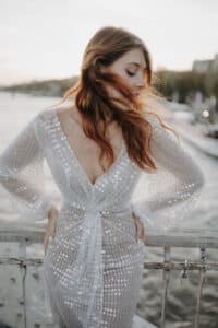 Eine Frau in einem glitzernden Paillettenkleid steht mit abgewandtem Gesicht auf einer Brücke, ihr Haar weht im Wind. Im Hintergrund sind ein Fluss und eine verschwommene Landschaft zu sehen, die eine ruhige und elegante Atmosphäre schaffen.