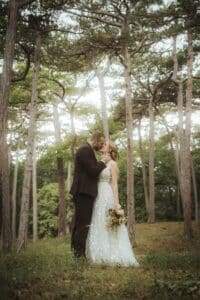 Braut und Bräutigam stehen liebevoll zwischen hohen Bäumen. Der Bräutigam im dunklen Anzug hält die Braut, die ein fließendes weißes Kleid trägt und einen Blumenstrauß hält, sanft im Arm. Sonnenlicht fällt durch die Zweige und schafft eine heitere und romantische Atmosphäre.