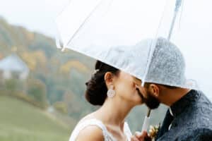 Braut und Bräutigam küssen sich an einem regnerischen Tag unter einem durchsichtigen Regenschirm. Die Braut trägt eine elegante Hochsteckfrisur und Ohrringe, während der Bräutigam einen Bart und formelle Kleidung trägt. Im Hintergrund sind verschwommenes Grün und ein angedeutetes Gebäude zu sehen.