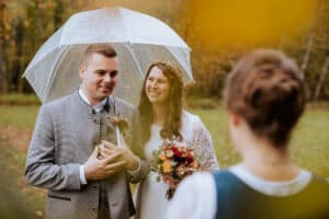 Braut und Bräutigam stehen lächelnd im Freien unter einem durchsichtigen Regenschirm. Der Bräutigam trägt einen grauen Anzug und die Braut hält einen Blumenstrauß. Sie blicken auf eine Person im Vordergrund, die ihnen den Rücken zukehrt. Es ist ein regnerischer Tag mit Herbstlaub.