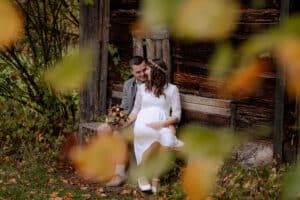 Ein Paar sitzt auf einer Holzbank vor einer rustikalen Hütte. Die Frau im weißen Kleid hält einen Blumenstrauß in der Hand, neben ihr steht der Mann. Herbstblätter rahmen die Szene ein und verleihen ihr einen Hauch von Wärme und Farbe.