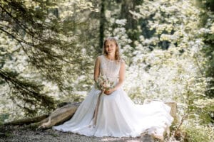 Eine Braut in einem weißen Kleid sitzt auf einem umgestürzten Baum in einem sonnenbeschienenen Wald, lächelt und hält einen Blumenstrauß. Der Hintergrund ist üppig grün und von gesprenkeltem Sonnenlicht durchflutet.