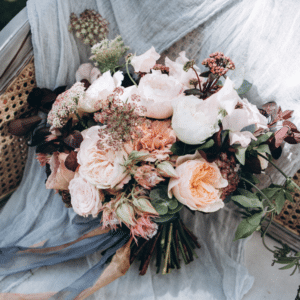 Ein Strauß pastellfarbener Blumen, darunter Rosen und Pfingstrosen, liegt auf einem hellblauen Stoff, der über einem Stuhl drapiert ist. Das Arrangement besteht aus zarten Rosa- und Cremetönen sowie dezentem Grün und schafft so ein romantisches und elegantes Arrangement.