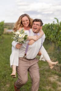 Ein fröhliches Paar im Hochzeitskleid steht in einem Weinberg. Die Frau wird von dem Mann spielerisch Huckepack getragen und hält einen Strauß weißer Blumen in der Hand. Umgeben von grünen Weinreben und einem bewölkten Himmel lächeln beide breit.