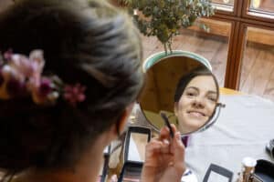 Eine Frau lächelt, während sie in einen runden Spiegel schaut und sich mit einem Pinsel schminkt. Sie sitzt an einem Tisch mit verschiedenen Make-up-Artikeln. Ihr Haar ist mit Blumen gestylt und im Hintergrund ist eine Topfpflanze zu sehen.