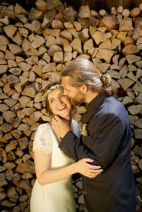 Ein Paar steht vor einem Stapel Brennholz. Der Mann im schwarzen Anzug küsst die Stirn der Frau. Sie trägt ein weißes Kleid mit Spitzenärmeln und einem Blumenkranz und lächelt freudig mit geschlossenen Augen.