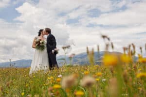 Braut und Bräutigam küssen sich auf einer malerischen Wiese voller bunter Wildblumen. Der Himmel ist teilweise bewölkt und im Hintergrund sind Berge zu sehen, was eine ruhige und romantische Atmosphäre schafft.