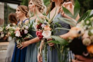 Frauen in farbenfrohen Kleidern halten Sträuße aus gemischten Blumen und Grünpflanzen und stehen bei einer Veranstaltung im Freien in einer Reihe. Der Fokus liegt auf den Blumenarrangements, mit verschwommenem Grün im Hintergrund.