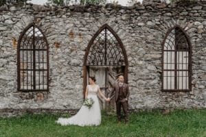 Eine Braut im weißen Kleid und ein Bräutigam im braunen Anzug stehen Händchen haltend vor einem alten Steingebäude mit Bogenfenstern. Die Braut hält einen Blumenstrauß. Die Szene hat eine rustikale, romantische Atmosphäre.